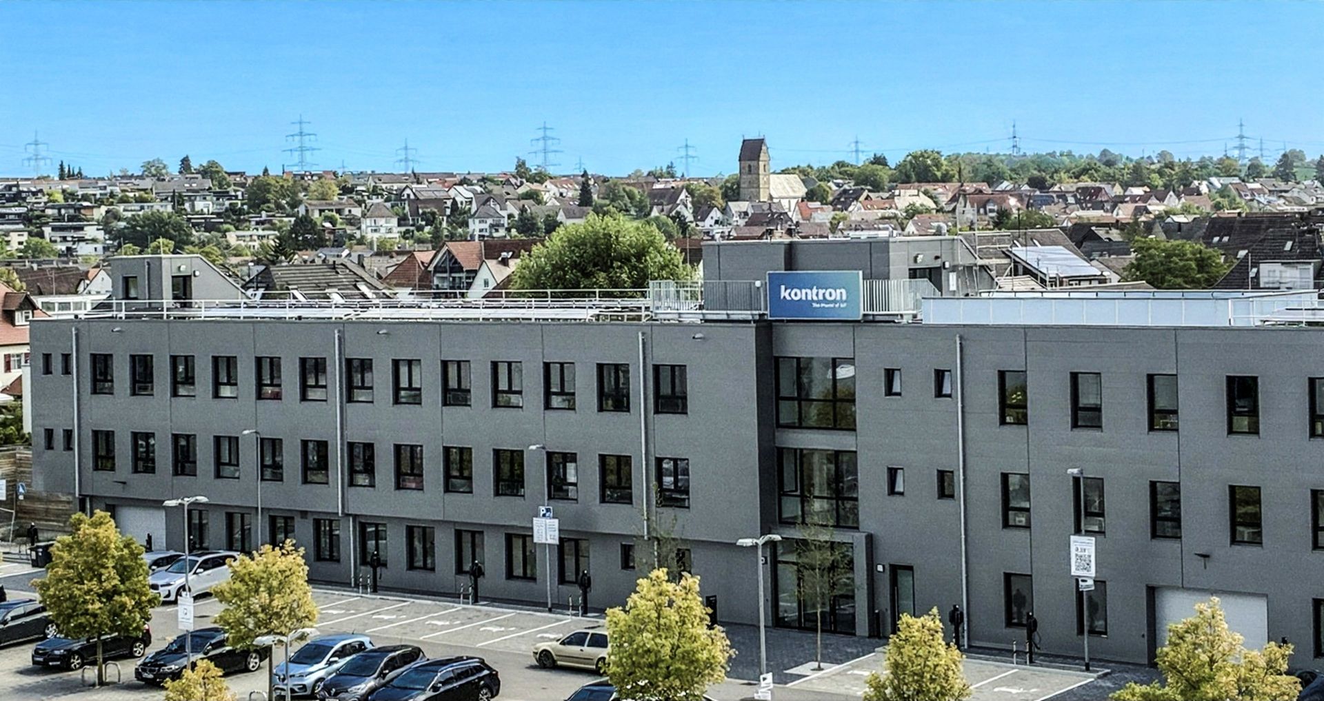 Moderne Bürogebäude mit großen Fenstern und einem Eingangsbereich. Vor dem Gebäude befinden sich parkende Autos und Bauarbeiten im Gange. Im Hintergrund sind Wohngebäude und eine ländliche Umgebung sichtbar.