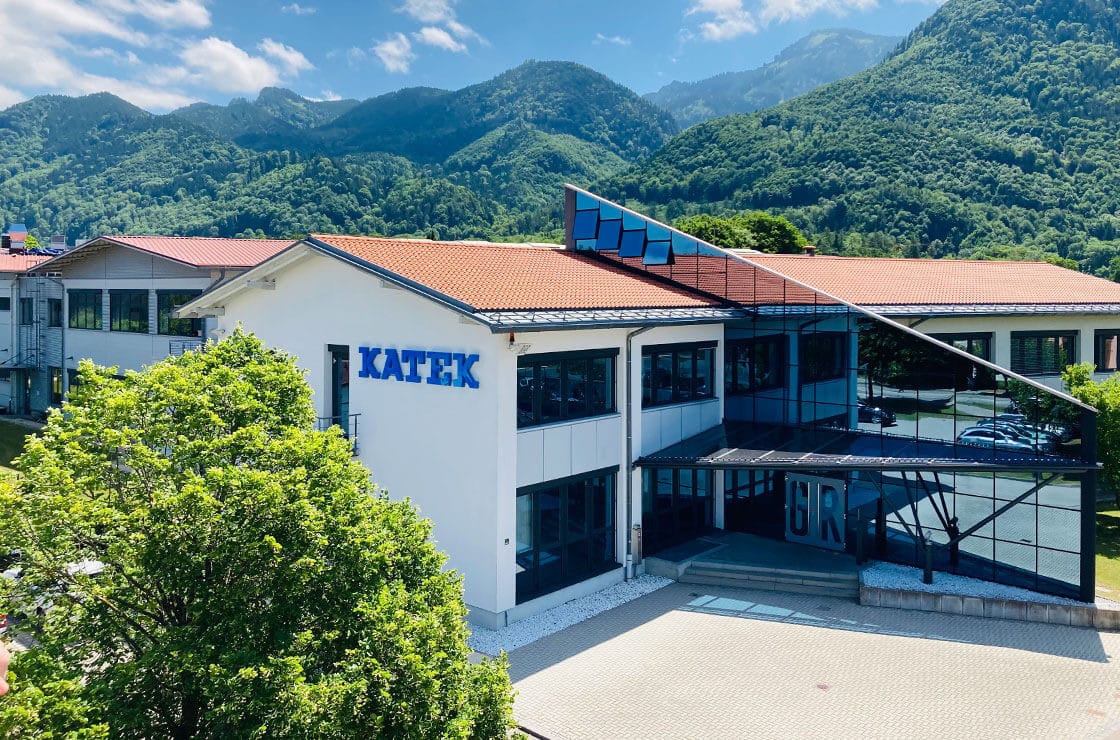 A modern building with the name "Katek" prominently displayed on the facade, surrounded by lush greenery and mountains in the background under a blue sky.
