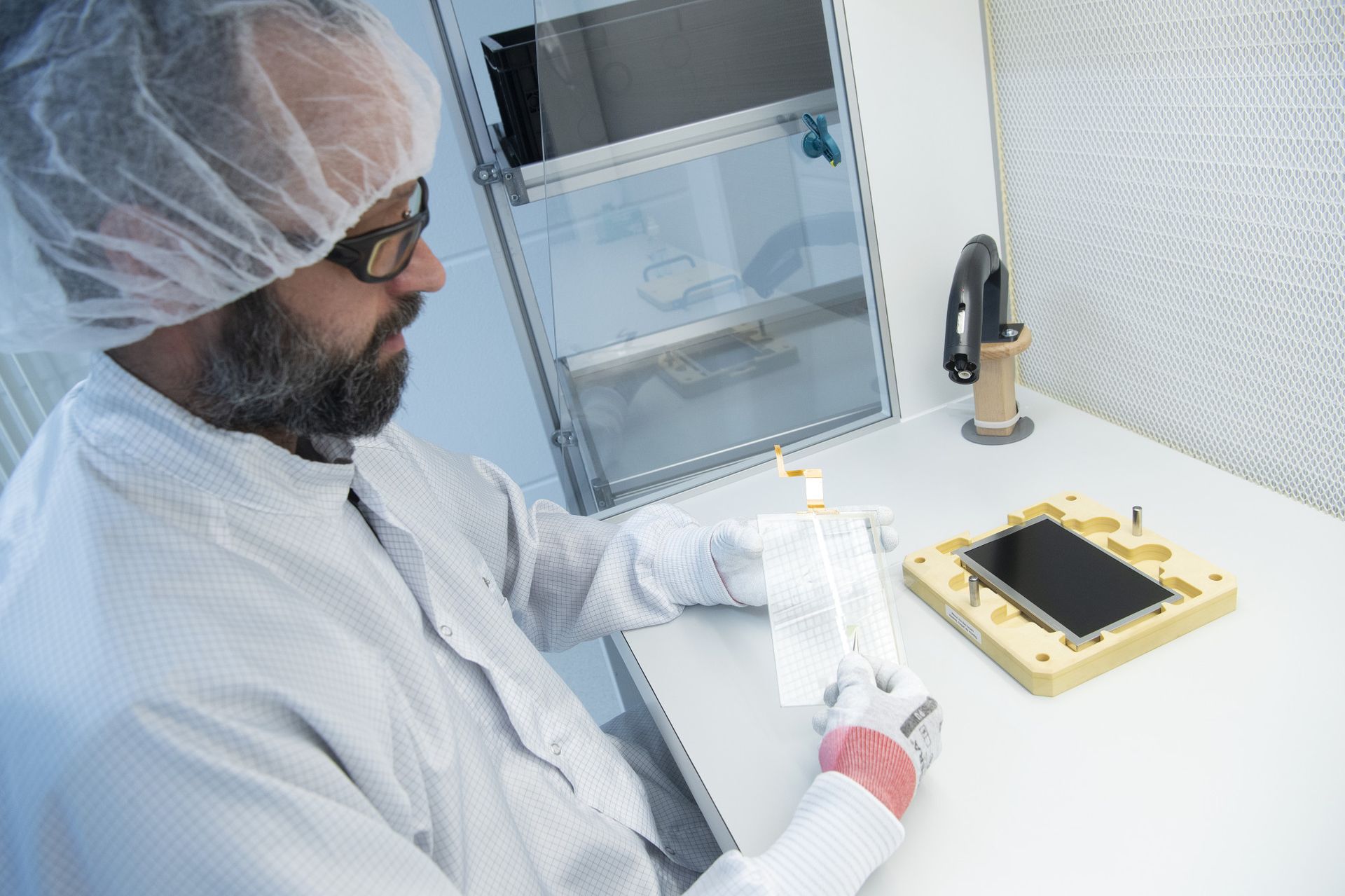 A researcher in a lab coat and hairnet examines a transparent slide in a cleanroom. A black device is positioned on a wooden platform nearby, with lab equipment and a protective enclosure in the background.