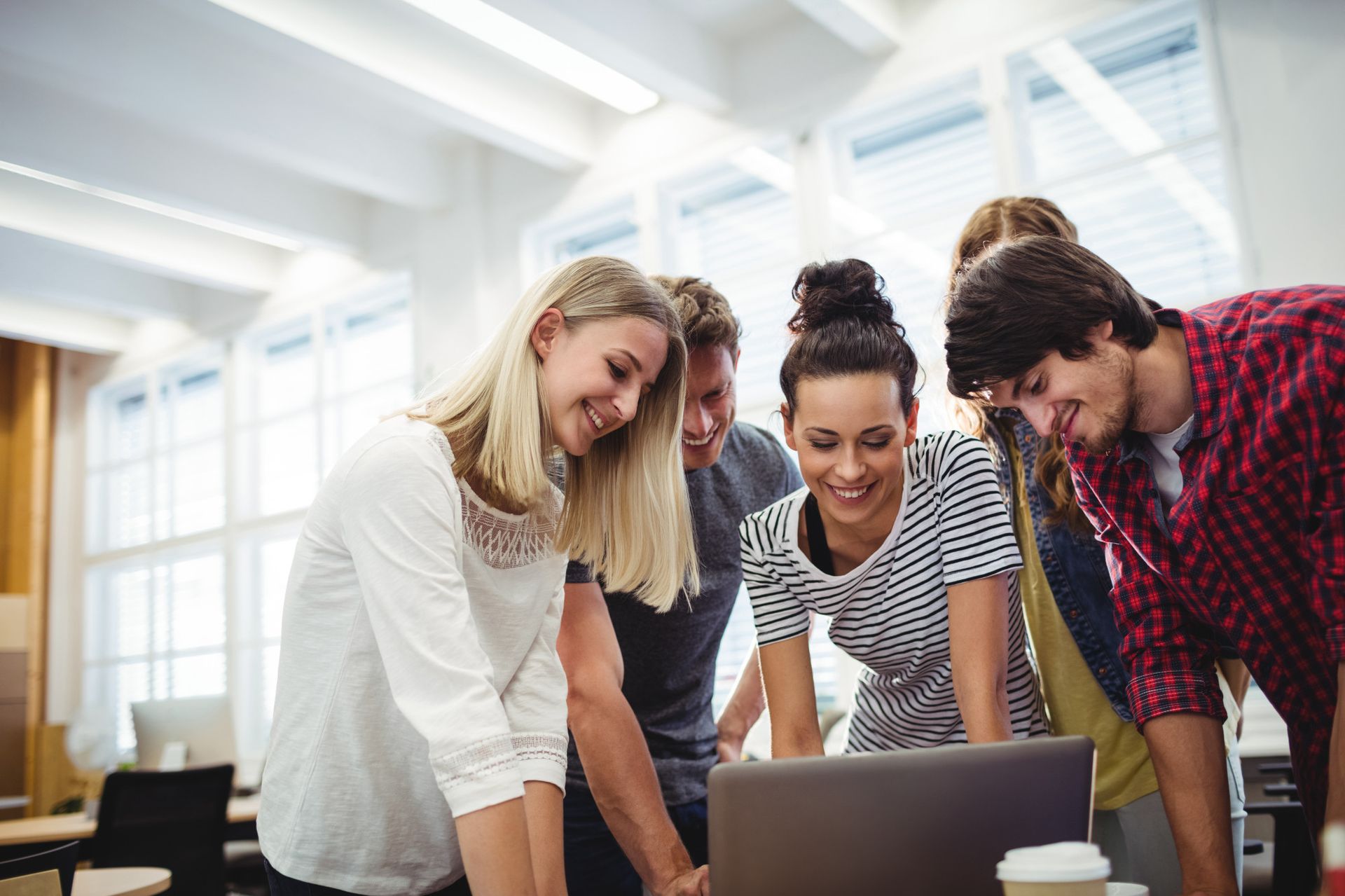 A group of five young adults gather around a laptop, smiling and engaged in discussion. They are in a bright, modern workspace with large windows and a relaxed atmosphere.