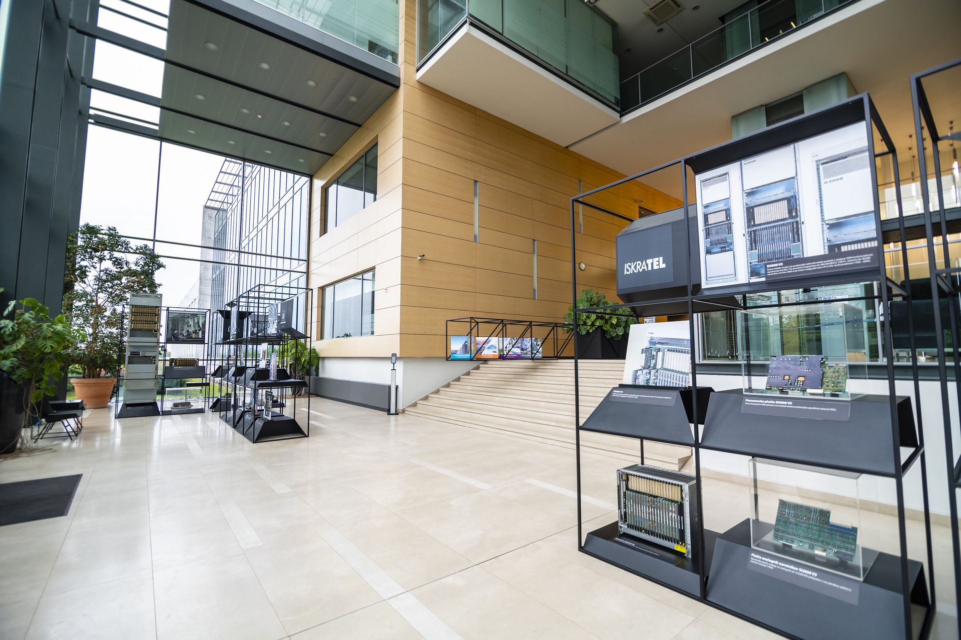 A spacious modern interior with large windows showcasing an exhibition. Black display stands feature various electronic circuit boards and models. The setting includes a staircase and greenery, emphasizing a sleek architectural design.