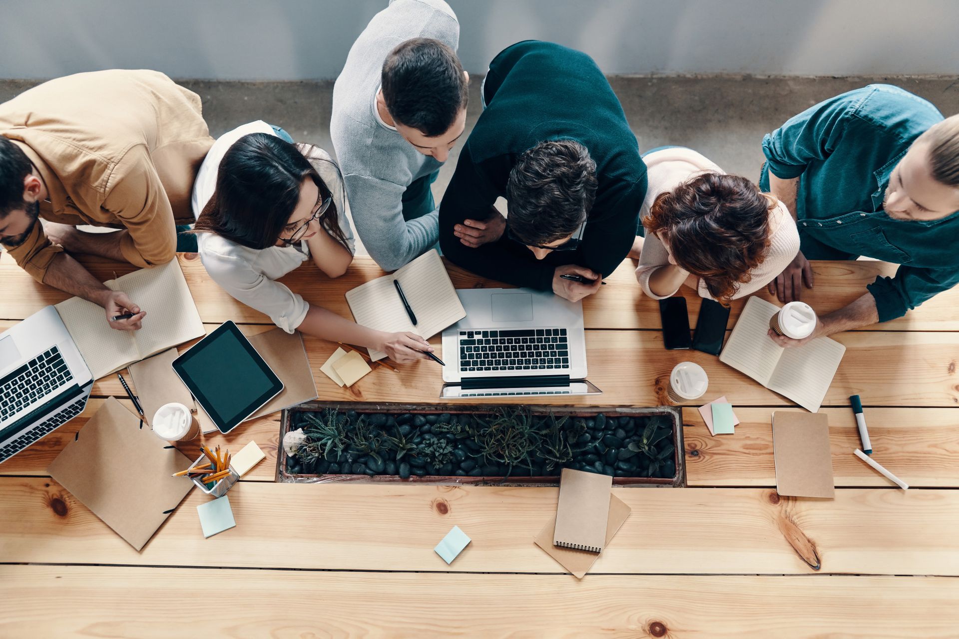 A group of people sitting around a wooden table, collaborating on a project. Laptops, notebooks, and coffee cups are scattered across the table, with a small planter featuring greenery in the center. They appear engaged and focused on a laptop screen.