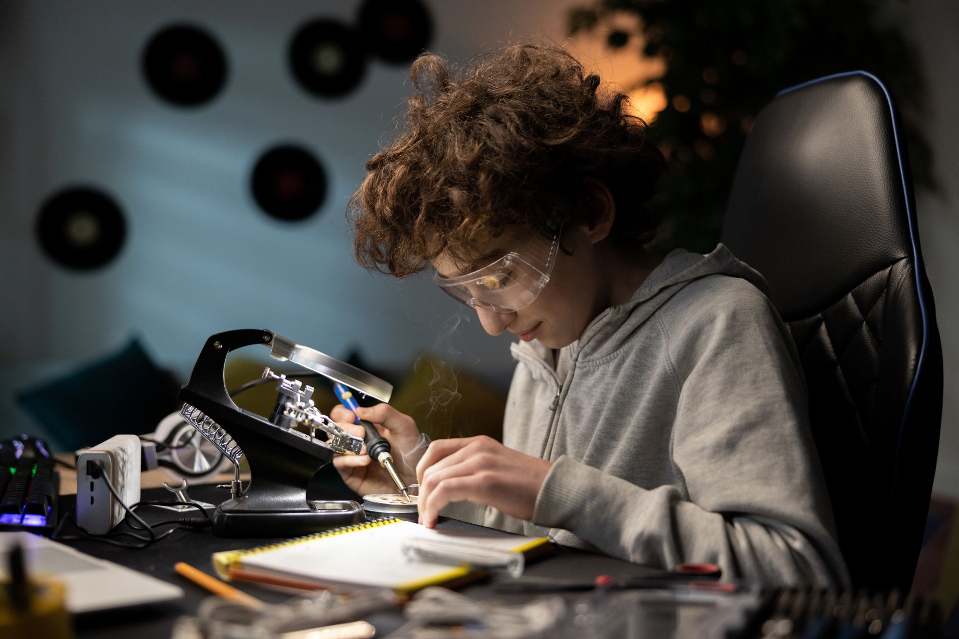 A young boy with curly hair and safety glasses focuses on soldering components at a desk. Tools and electronic parts are scattered around, and a notepad is open beside him.