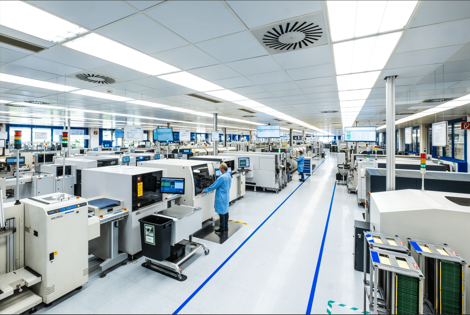 Große, moderne Fabrikhalle mit mehreren Produktionslinien. Mitarbeiter arbeiten an Maschinen, Bildschirme zeigen technische Informationen. Helle Beleuchtung und klare Bodenmarkierungen sind sichtbar.