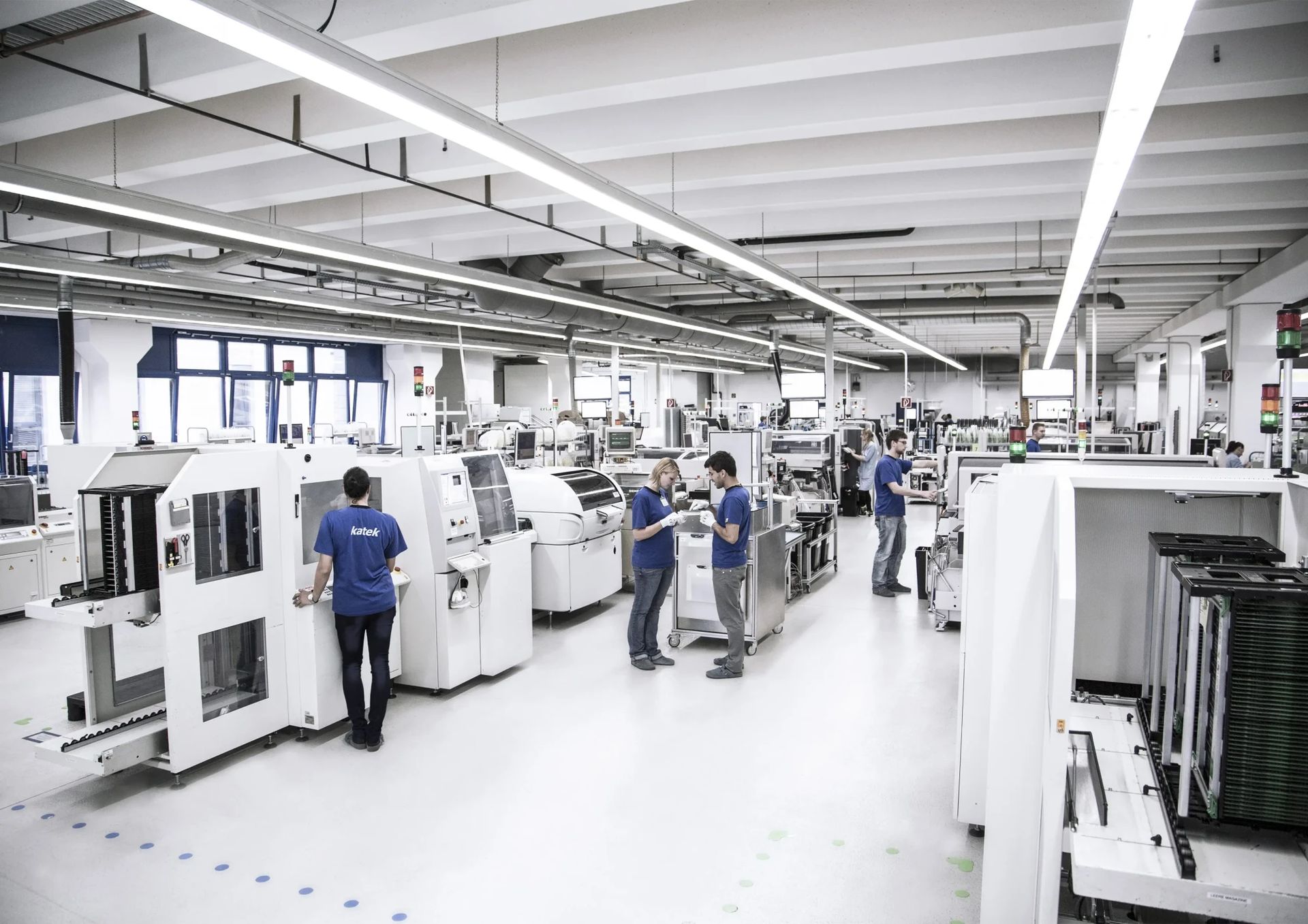 A large, modern industrial facility filled with machines and workers. Several employees in blue shirts are operating equipment and moving between machines in a spacious, well-lit environment.