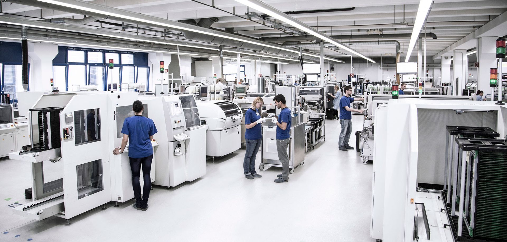 A spacious manufacturing facility with multiple machines and equipment. Workers in blue shirts are engaged in tasks around the machines, which are mostly white and industrial in design. Bright overhead lighting illuminates the area, ensuring clear visibility.
