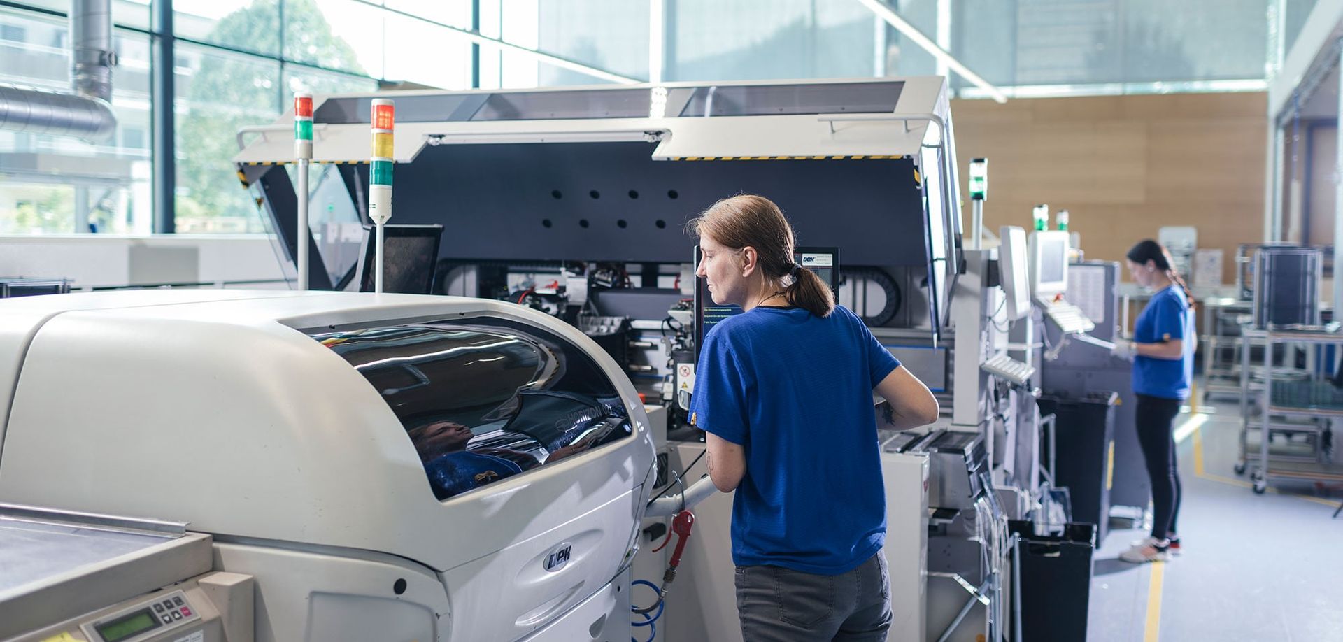 A woman in a blue shirt operates a large machine in a modern manufacturing facility, while another employee works at a nearby station. The bright, spacious environment features large windows and high-tech equipment.
