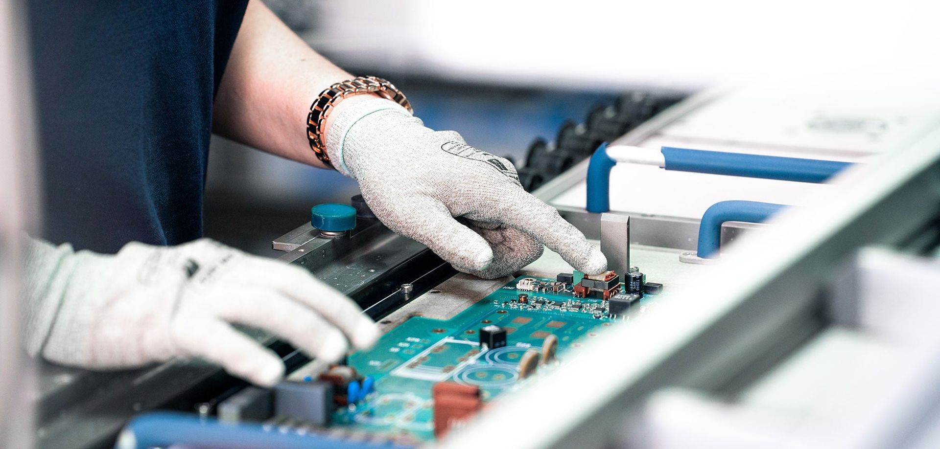 A person in gloves is working on a printed circuit board, carefully touching a component while focusing on the details. The work area is equipped with machinery and tools.