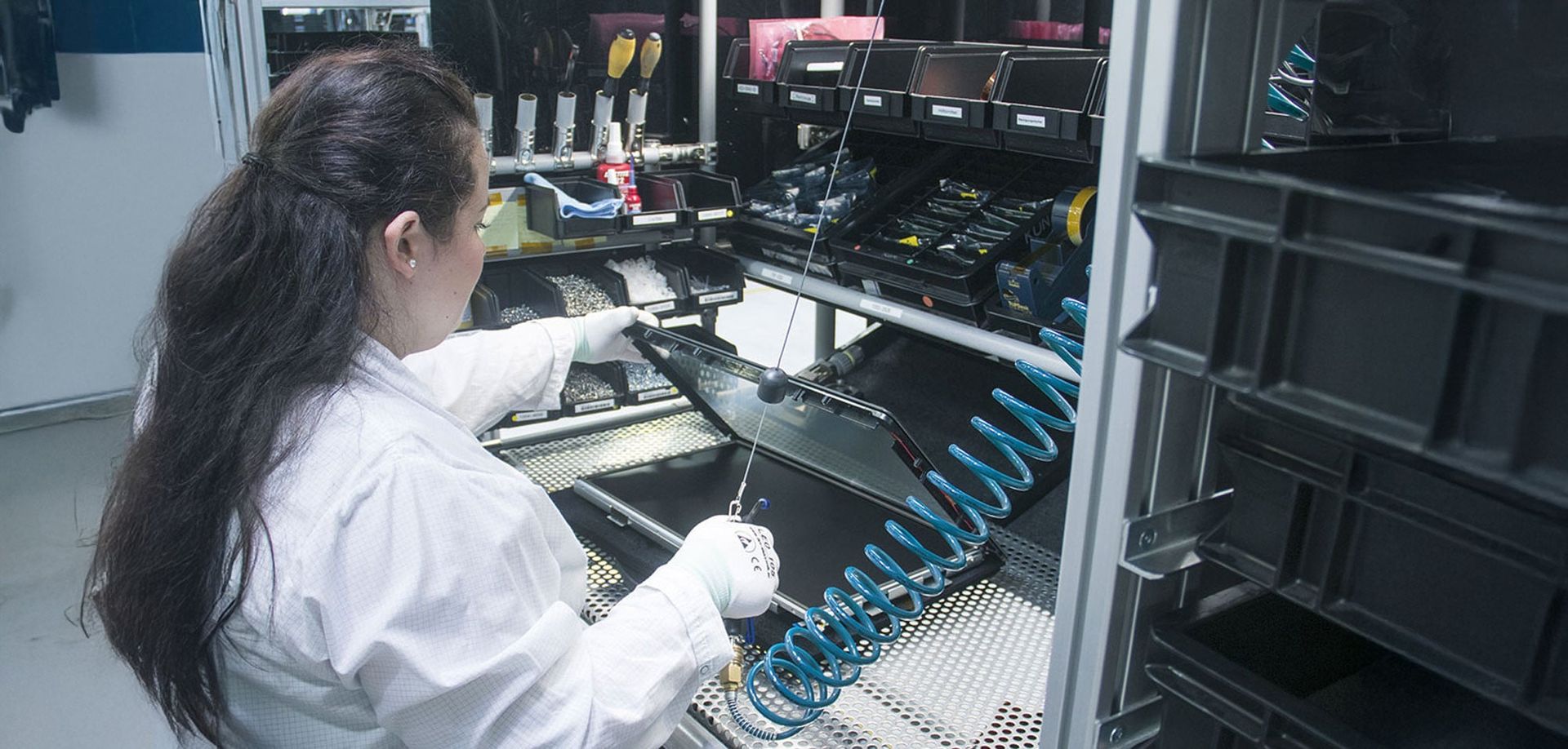 A technician in a lab coat works on a sleek device at a workstation, using tools and equipment organized in a shelf. The environment is clean and industrial, with various instruments and cables visible.