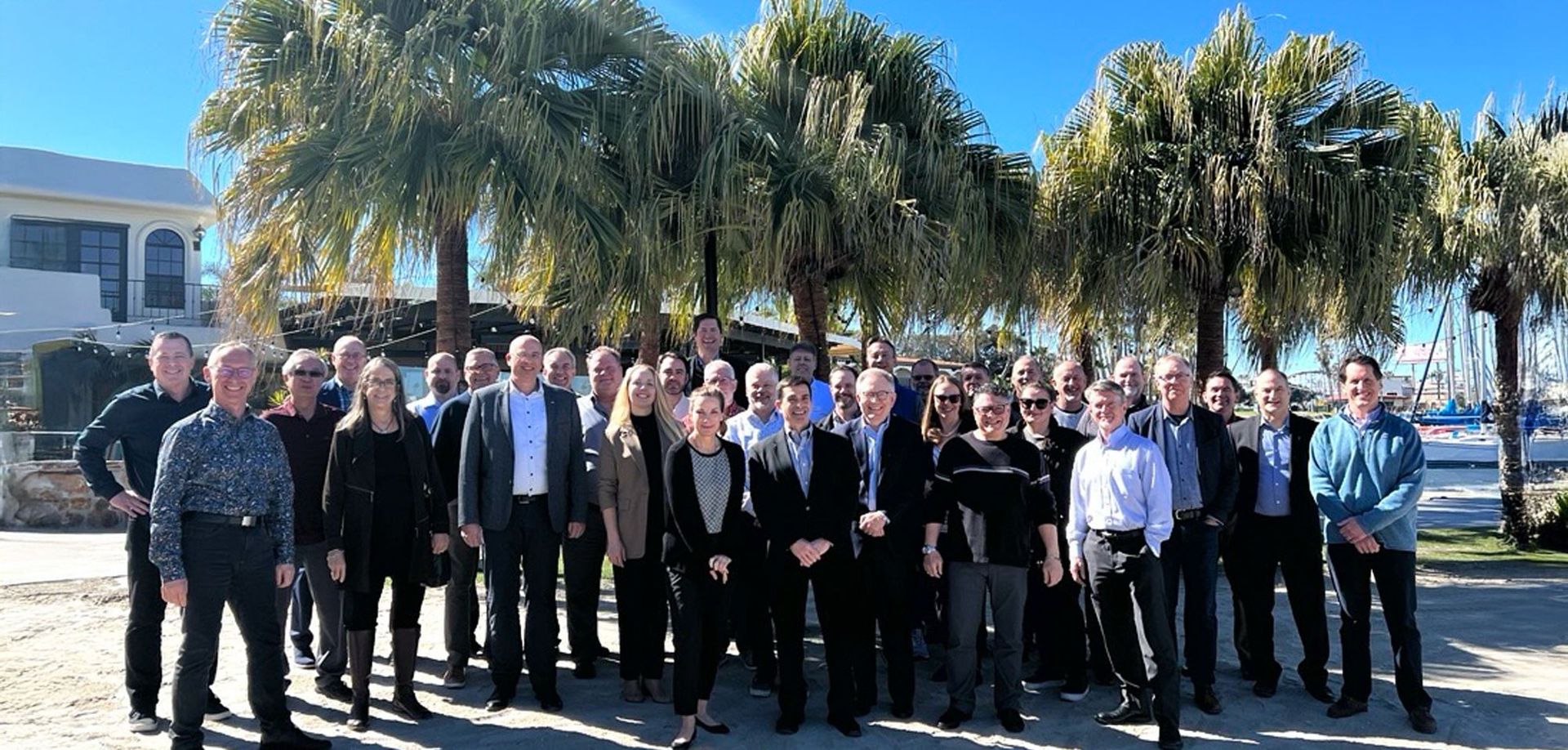 A diverse group of people stands outside under palm trees, smiling and posing for a photo. The scene features a sunny sky with a marina in the background.