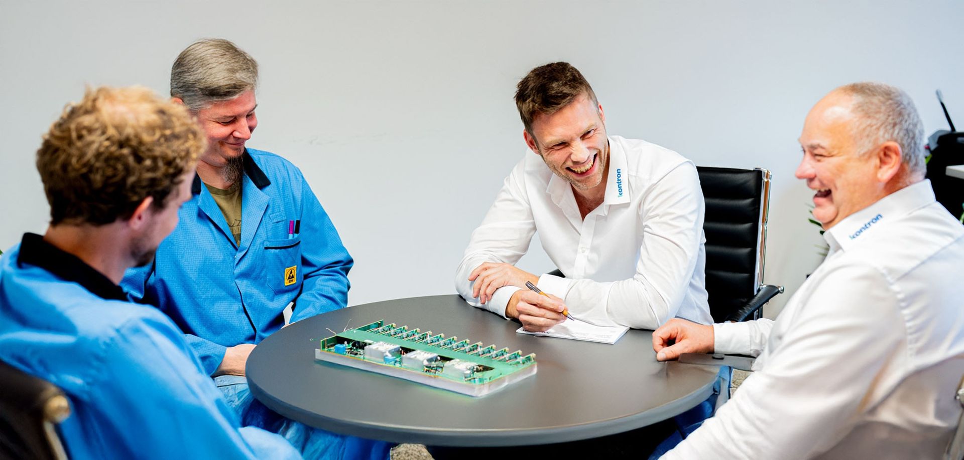 Vier Personen sitzen an einem runden Tisch und diskutieren. Zwei tragen blaue Overalls, während einer ein weißes Hemd und der andere ein graues Hemd trägt. Auf dem Tisch liegt eine Leiterplatte. Alle lachen und scheinen engagiert.