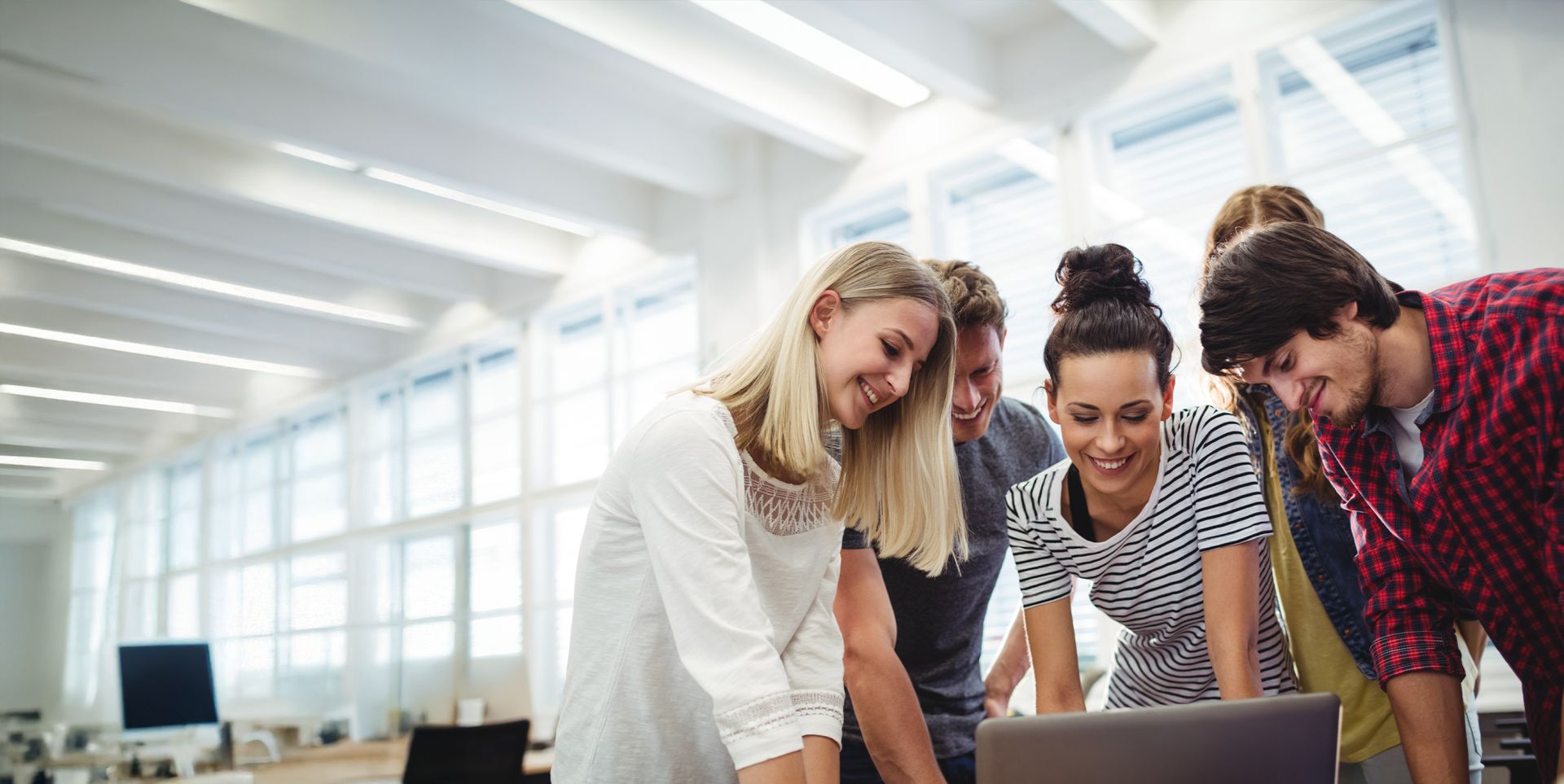 A group of five young people gathered around a laptop, smiling and engaging with each other in a bright, modern office environment with large windows.