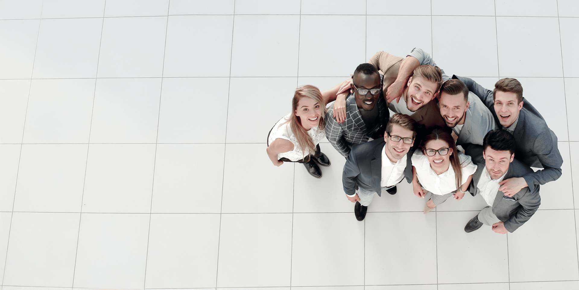A group of nine young adults smiles at the camera, standing closely together on a white tiled floor. They are dressed in a mix of business casual and formal attire.