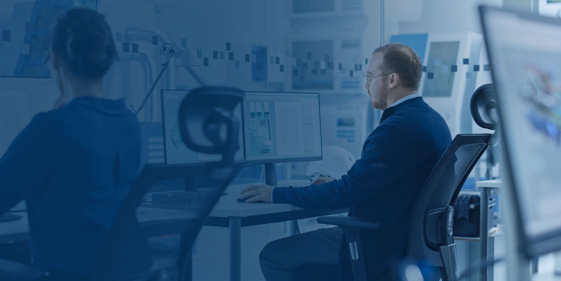 Ein Mann sitzt an einem Schreibtisch vor zwei Monitoren und arbeitet konzentriert. Im Hintergrund ist eine Person zu sehen, die ebenfalls am Computer arbeitet. Die Szene ist in Blautönen gehalten.