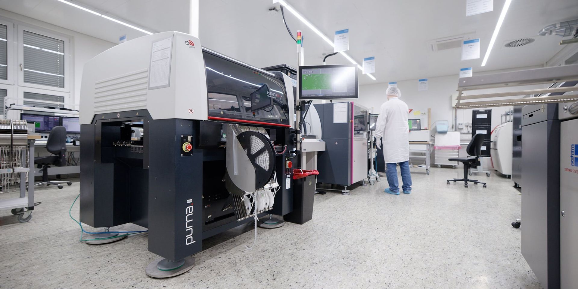 A modern manufacturing facility featuring advanced machinery, including a pick-and-place machine labeled "Puma II." An individual in a white lab coat and hairnet is visible in the background, working at a workstation with various equipment and screens. The environment appears clean and organized.