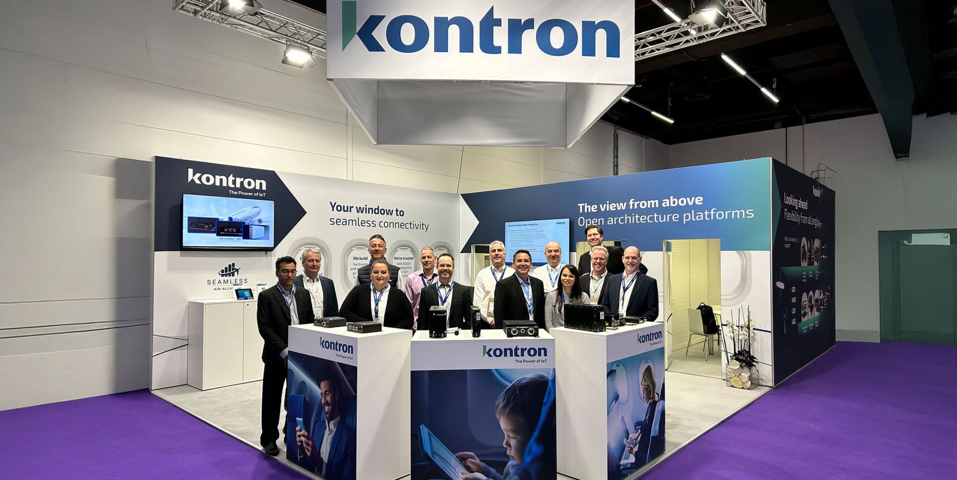 Un groupe de personnes pose devant un stand d'exposition de la société Kontron, avec des affiches sur la connectivité et des plateformes d'architecture ouverte.