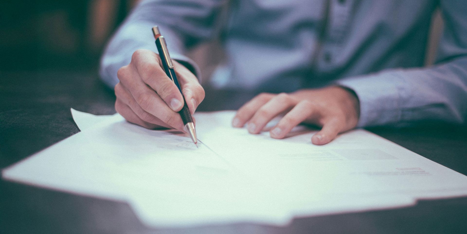 A person's hand holding a pen, writing on sheets of paper on a flat surface. The individual is wearing a light blue shirt.