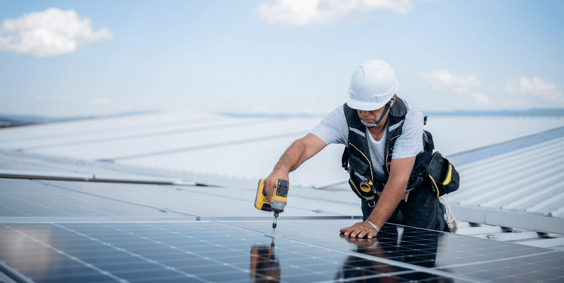 Ein Arbeiter mit Helm und Werkzeugtasche installiert Solarpaneele auf einem Dach. Er verwendet einen Schraubendreher, während er sich auf die Paneele konzentriert. Der Himmel ist blau mit einigen Wolken.