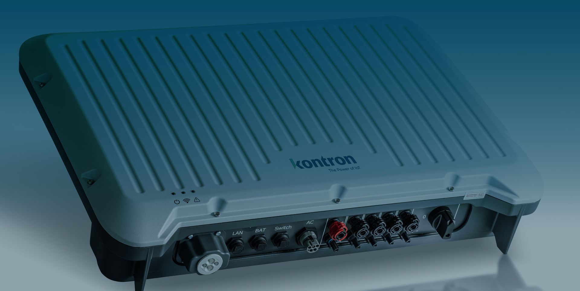 Ein graues, rechteckiges Gerät mit dem Schriftzug "kontron" auf der Oberseite. An der Vorderseite befinden sich verschiedene Anschlüsse für LAN, Batterien, Schalter und Stromversorgung. Die Oberflächenstruktur ist gerippt und das Design ist modern.