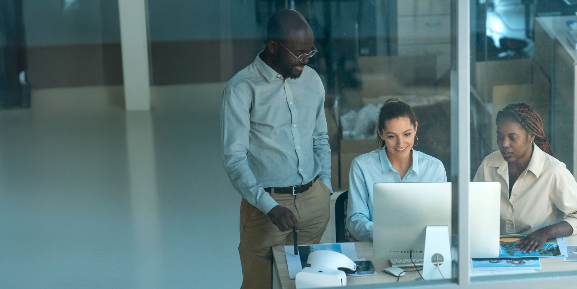 A group of three people are collaborating at a desk with a computer. Two women and one man are engaged in a discussion, with the man standing and the women seated. They appear focused and are looking at the computer screen, surrounded by a modern office environment.