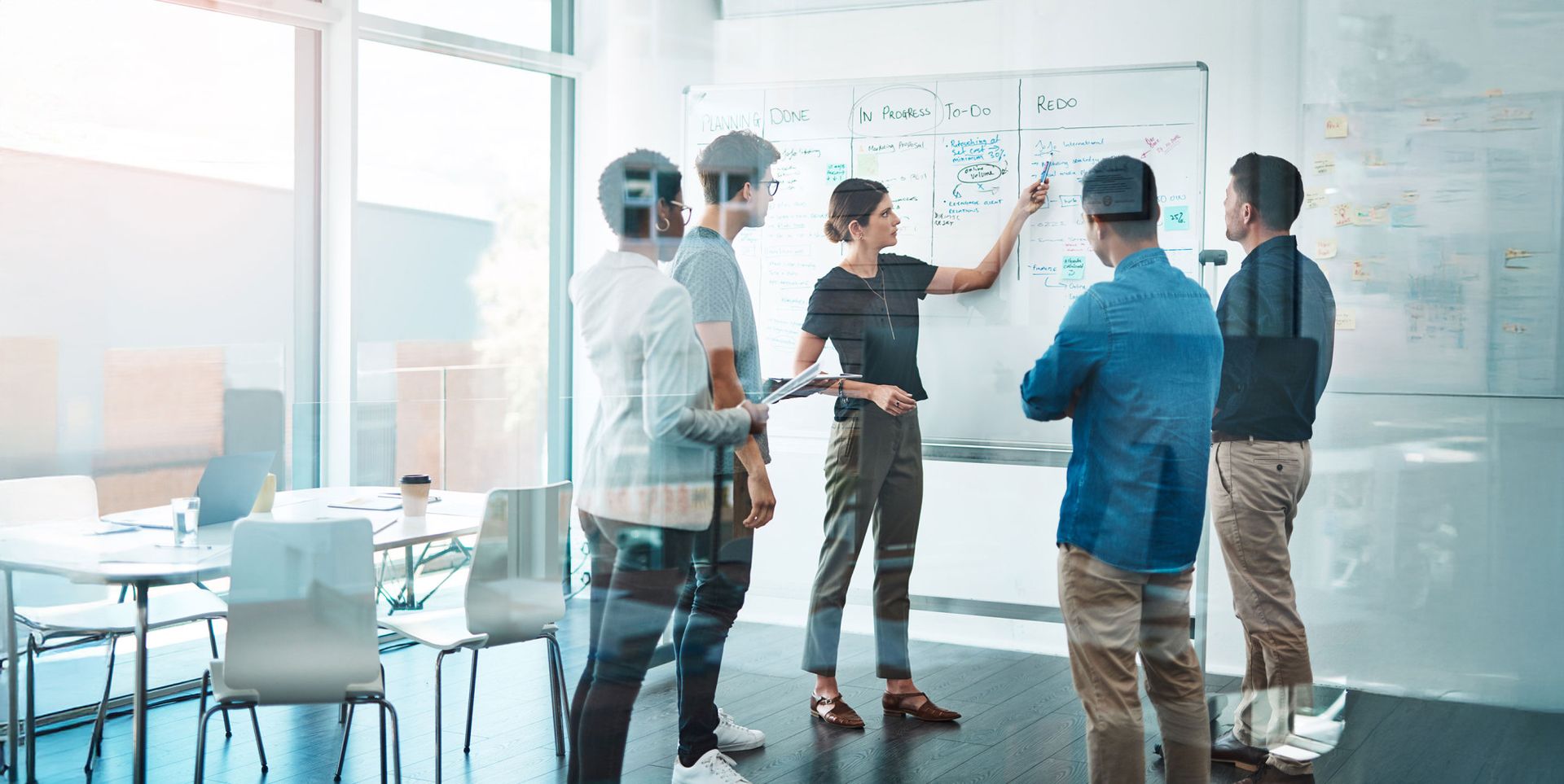 A group of six people engages in a discussion while standing around a whiteboard covered with notes and diagrams. The setting is a modern office with large windows and a collaborative atmosphere.