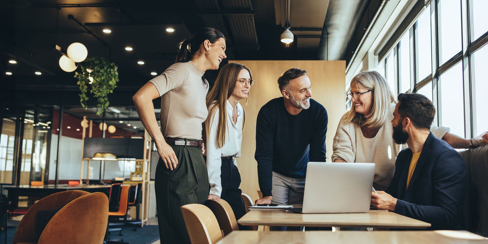 A group of five professionals, engaged in discussion around a laptop, are gathered in a modern office space. They appear to be collaborating, with smiles and focused expressions, while seated at a wooden table. Natural light filters in through large windows, creating a vibrant atmosphere.