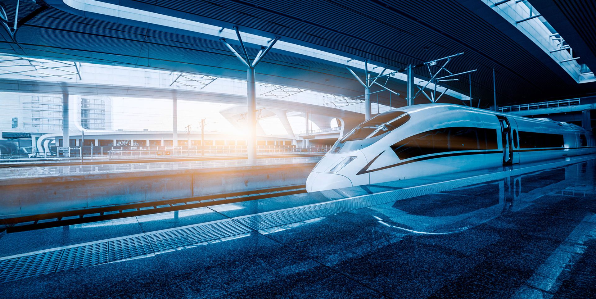 A modern high-speed train is parked at a sleek, contemporary train station. Bright sunlight streams through the large windows, reflecting off the polished floor surfaces. The architecture features clean lines and an open design.