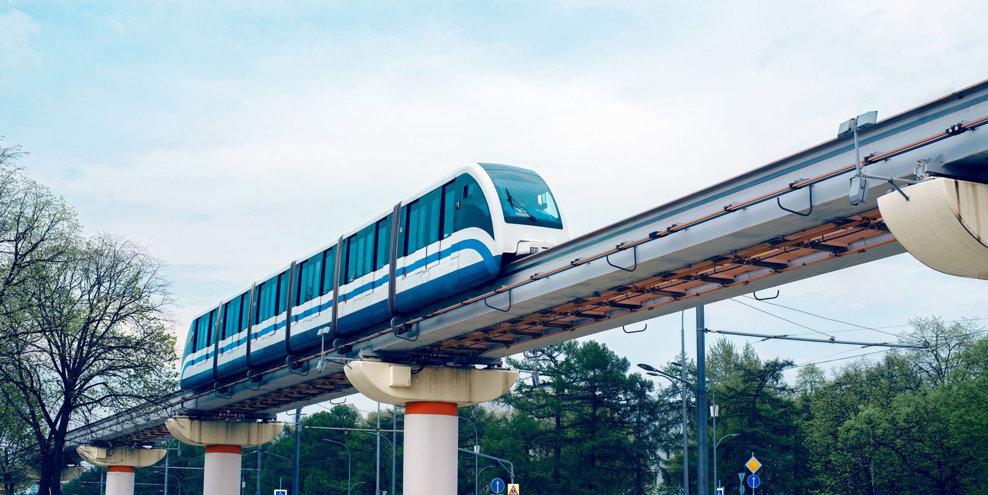 A modern monorail train glides above ground on a concrete track, surrounded by green trees and a cloudy sky.