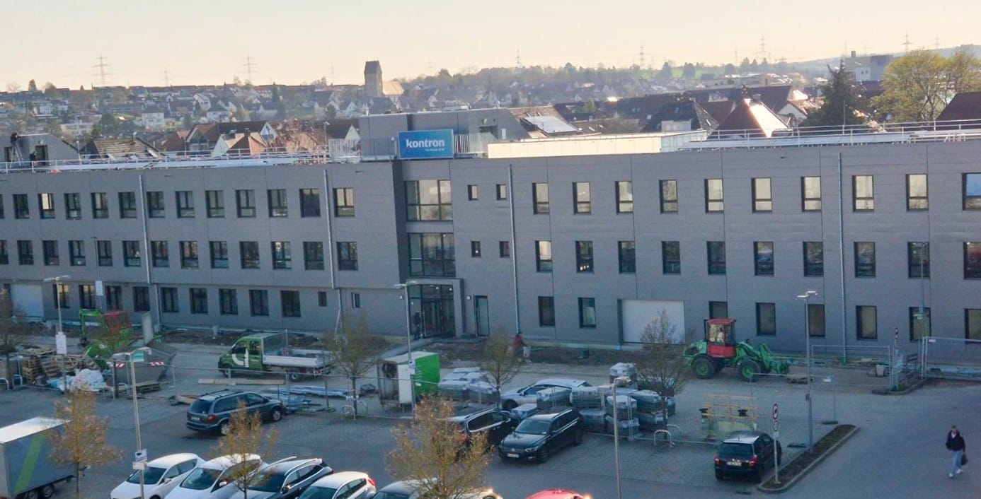 Moderne Bürogebäude mit großen Fenstern und einem Eingangsbereich. Vor dem Gebäude befinden sich parkende Autos und Bauarbeiten im Gange. Im Hintergrund sind Wohngebäude und eine ländliche Umgebung sichtbar.