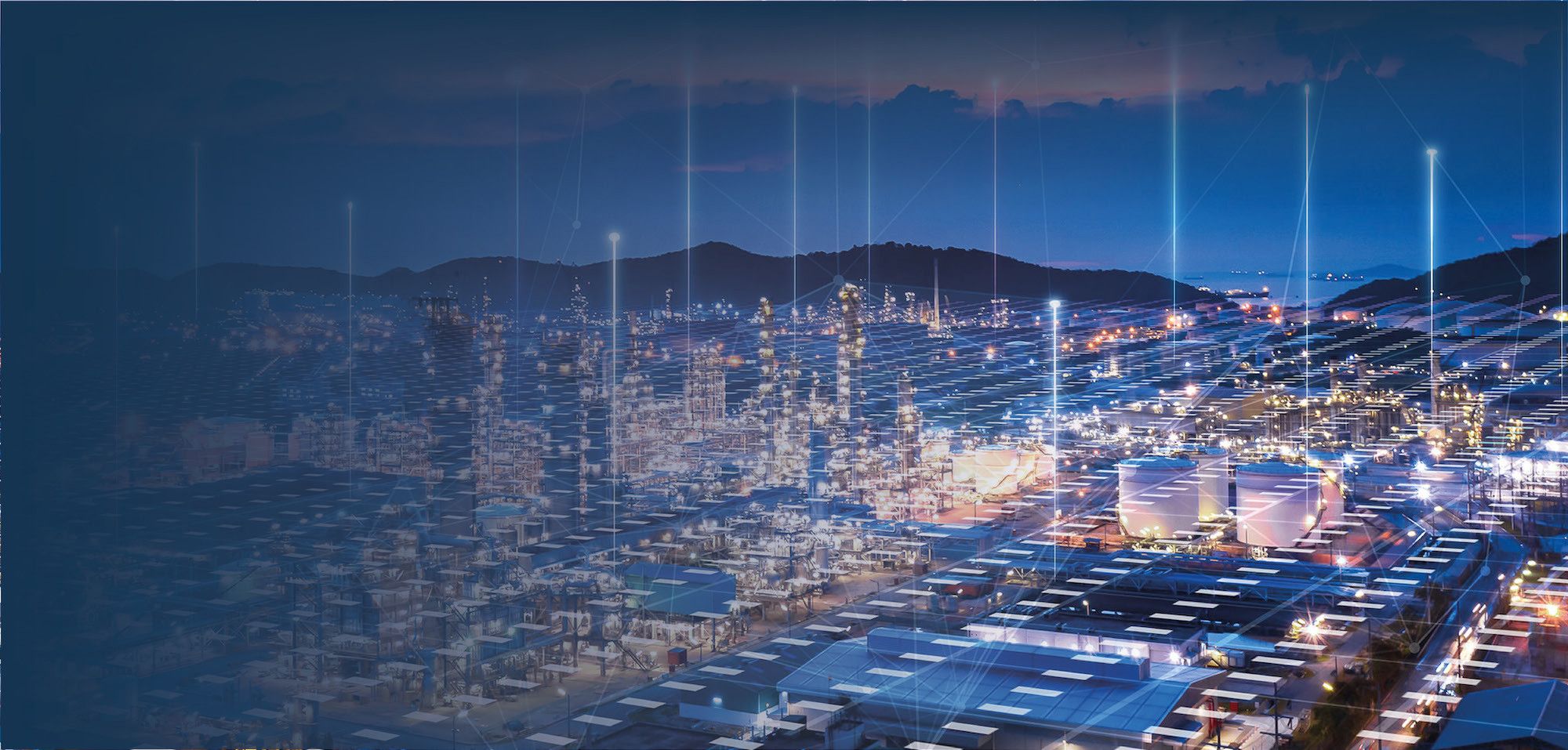 Eine moderne Stadtlandschaft bei Nacht, beleuchtet von zahlreichen Lichtern. Hochhäuser, Lagerhäuser und Straßen sind sichtbar, während ein futuristisches Netzwerk von Datenströmen durch die Luft visualisiert wird. Im Hintergrund sind sanfte Hügel und ein Gewässer zu sehen.