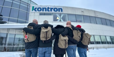 Un groupe de personnes de dos, avec des sacs à dos, se tenant par les épaules devant un bâtiment portant le logo "Kontron" sous un ciel neigeux.