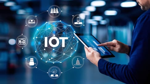 Eine Person hält ein Tablet und interagiert mit einem stilisierten Diagramm, das das Internet der Dinge (IoT) zeigt. Um das zentrale IoT-Symbol sind verschiedene Icons für vernetzte Geräte angeordnet.