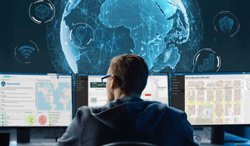 A person in a hoodie sits at a desk with multiple monitors displaying maps, data analytics, and cybersecurity icons. A large, illuminated globe with network lines hovers in the background, symbolizing global connectivity and data monitoring.