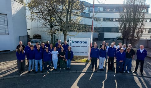 A large group of employees stands in front of a sign for Kontron, wearing matching blue shirts, with a modern building in the background. Trees are visible, indicating a sunny day.