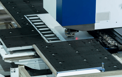 A CNC machine operates on a workpiece, with a laser marking tool visible. The machine features a conveyor system with textured black surfaces and multiple holes for securing the material.