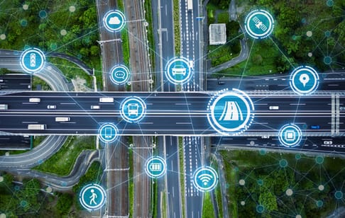 An aerial view of a highway intersection with digital icons representing various transportation and connectivity features, including public transport, GPS, and smart technology. Green trees surround the area, indicating a mix of urban and natural elements.