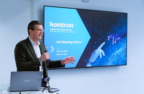 A man stands at a podium, holding a microphone while speaking during a presentation. A large screen behind him displays the title "SLA Meeting Vienna" along with the date and the Kontron Group logo. There is a digital image of a robotic hand interacting with a holographic interface on the screen.