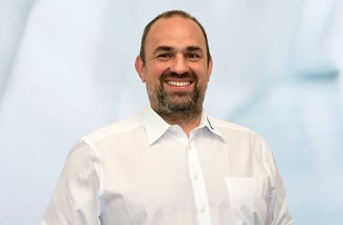 Ein lächelnder Mann mit Kurzhaarschnitt und Vollbart trägt ein weißes Hemd. Der Hintergrund ist unscharf in blauen und weißen Tönen.