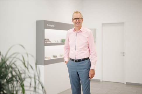 Eine lächelnde Person in einem hellen, modernen Raum. Er trägt ein hellrosa Hemd und blaue Hose und steht entspannt neben einem Regal mit Präsentationsmaterial. Im Hintergrund sind Pflanzen und eine weiße Wand zu sehen.