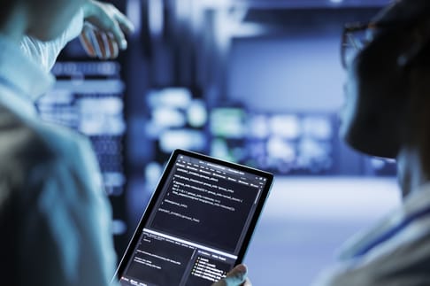 A person holding a tablet displaying code while another individual gestures towards interactive screens in a technology-driven environment. The background features blurred digital displays, creating a high-tech atmosphere.