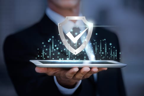 A businessman in a suit holds a tablet displaying a glowing shield with a checkmark, symbolizing digital security and protection.