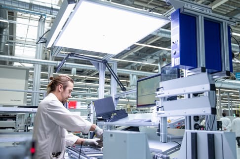 A technician works diligently at a workstation in a modern manufacturing facility, surrounded by equipment and a bright overhead light.