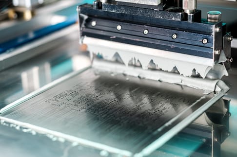 A close-up view of a printing squeegee, spreading ink across a screen printing frame. The surface shows a textured pattern where the ink is being applied.