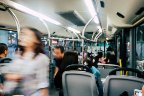 A crowded bus interior with passengers seated and standing. Some individuals are in motion, creating a blurred effect. The overhead lights illuminate the space, while the view outside the windows is partially visible.