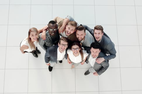 A group of nine smiling people stands in a circle, looking up at the camera. They are dressed in formal attire and appear to be enjoying a moment together in a bright, spacious environment with a tiled floor.