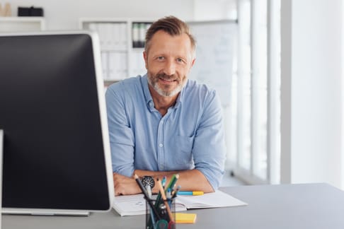 Ein lächelnder Mann mit Bart sitzt an einem Schreibtisch, vor einem Computerbildschirm. Er trägt ein hellblaues Hemd und hat Schreibutensilien sowie Unterlagen auf dem Tisch.