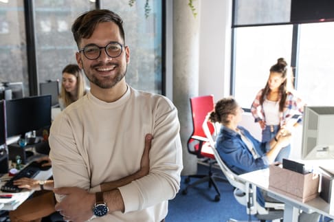 Ein junger Mann mit Brille und lächelndem Gesicht steht in einem modernen Büro. Im Hintergrund sind zwei Frauen zu sehen, die miteinander sprechen, während eine weitere Person am Schreibtisch arbeitet. Helle Fenster und eine freundliche Atmosphäre schaffen ein produktives Arbeitsumfeld.