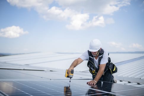 Ein Handwerker installiert eine Solaranlage auf dem Dach. Er trägt einen Helm und benutzt eine Bohrmaschine, um die Solarpaneele zu befestigen. Der Himmel ist blau mit vereinzelten Wolken.