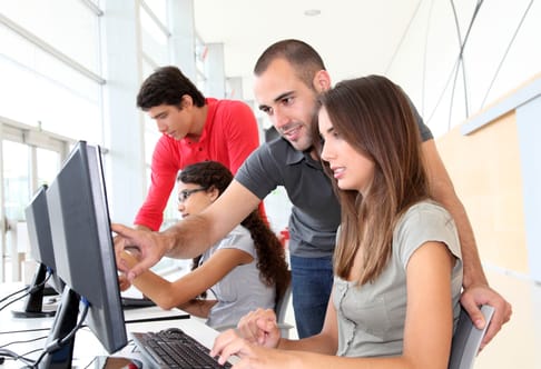 Vier Personen arbeiten in einem modernen Büro. Eine Frau sitzt am Computer und tippt, während ein Mann neben ihr auf den Bildschirm zeigt. Zwei weitere Personen sitzen im Hintergrund und sind ebenfalls mit ihren Computern beschäftigt. Helles, freundliches Umfeld mit großen Fenstern.