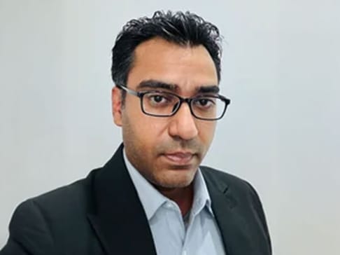 A man with short black hair and glasses poses for a selfie, wearing a black suit jacket over a light blue shirt. The background is plain and light-colored.